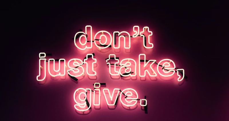Neonlicht mit der Botschaft 'don't just take, give.' in pinker Schrift auf dunklem Hintergrund.